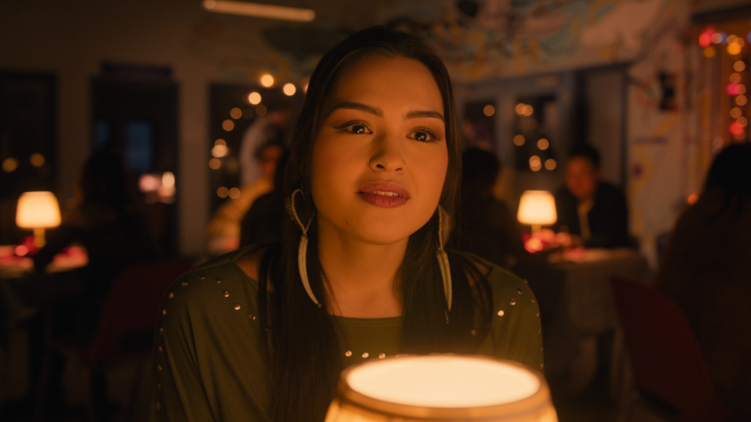 "Woman wearing feather earrings sits at a warmly lit table in a cozy, softly decorated restaurant with string lights, looking forward with a gentle expression. Other diners and ambient lamps are visible in the background."